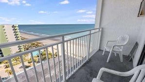 A balcony with a view of the beach and ocean. There are two white plastic chairs and a white railing.