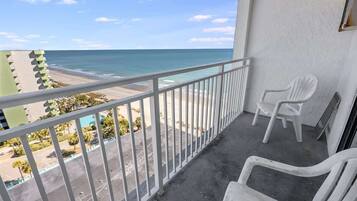 A balcony with a view of the beach and ocean. There are two white plastic chairs and a white railing.