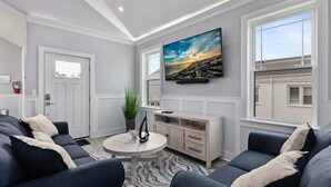 A modern living room with a sofa set, a coffee table, a TV mounted on the wall, and a window with a view of a building.