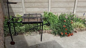A metal table with a grid top, positioned on a concrete surface, adjacent to a brick wall and a garden with various plants and flowers.