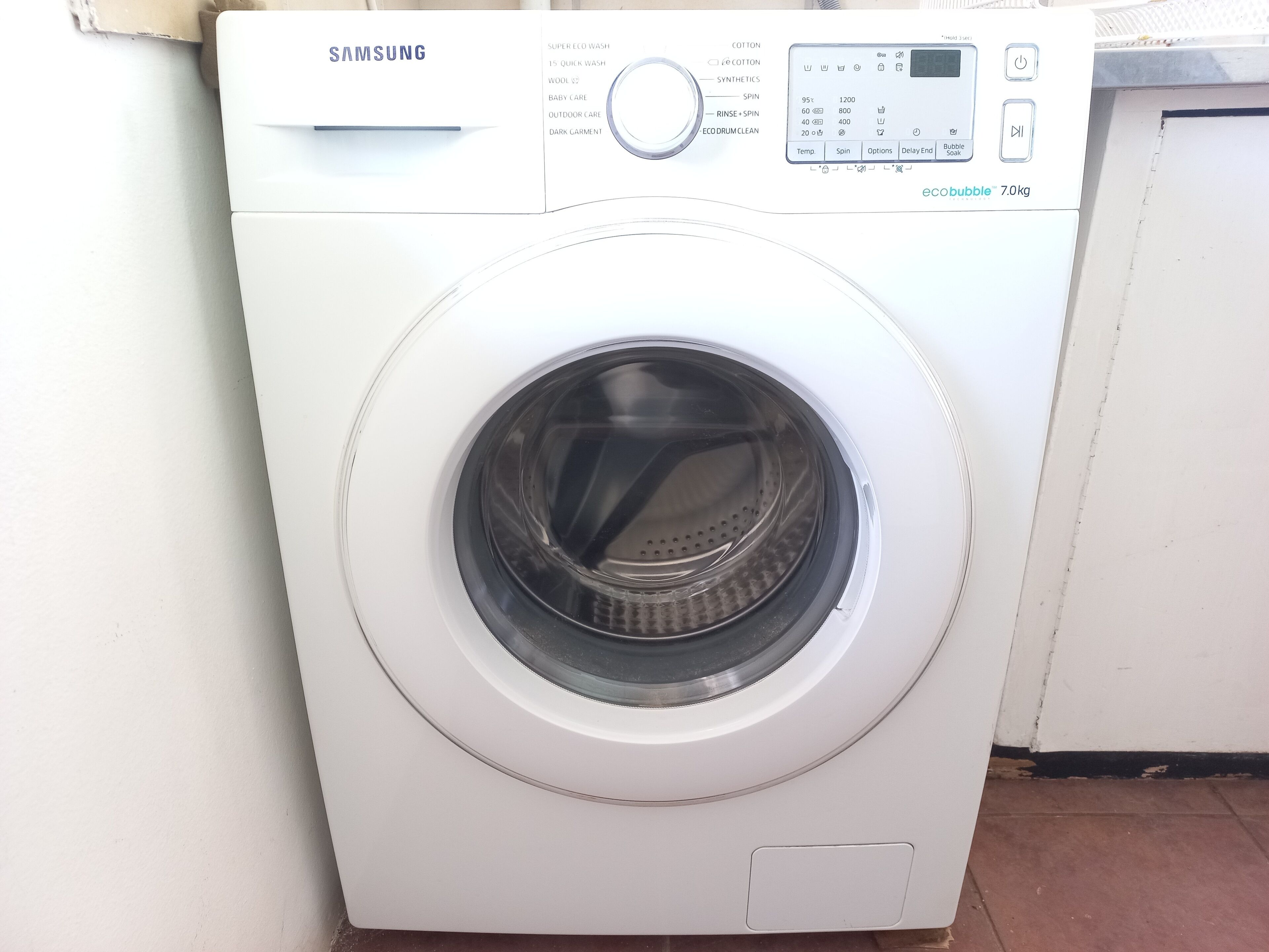 A Samsung washing machine with a control panel, a circular door, and a small storage compartment at the bottom.