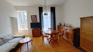 A living room with a sofa, a TV on a wooden stand, a dining table with chairs, and a kitchen area with cabinets and a refrigerator. - APPT F2 RDJ 4/5 PERSONS- ALL COMFORT - QUIET - CLOSE TO THERMAL BATHS  (La Bourboule)