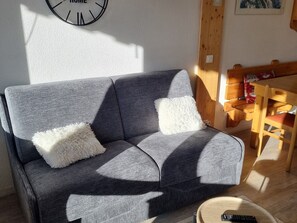 En grå soffa med vita, fluffiga kuddar, en trästol, ett runt träbord och en väggklocka.