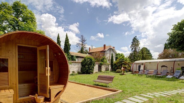 A wooden sauna structure in a garden with a stone pathway, a bench, and a small table with a bowl.