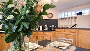 A dining area with a table set for two, a vase with flowers, a coffee machine, and a clock on the wall.