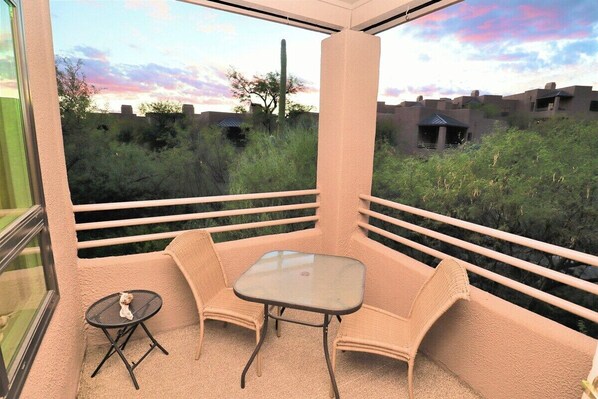 A balcony with wicker furniture and a glass table, overlooking a natural landscape with cacti and distant buildings.