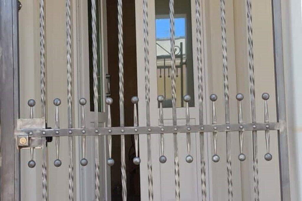 Une grille métallique avec des barreaux verticaux et des pommeaux sphériques, qui donne probablement accès à l’entrée d’un bâtiment.