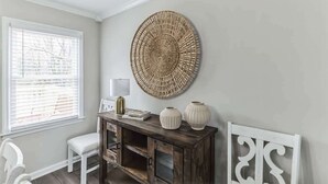 A room with a white chair, a wooden console table with decorative items, a round woven wall hanging, and a window with blinds. - Charming 3BR Farmhouse • Fenced Acre • Madison (Madison)