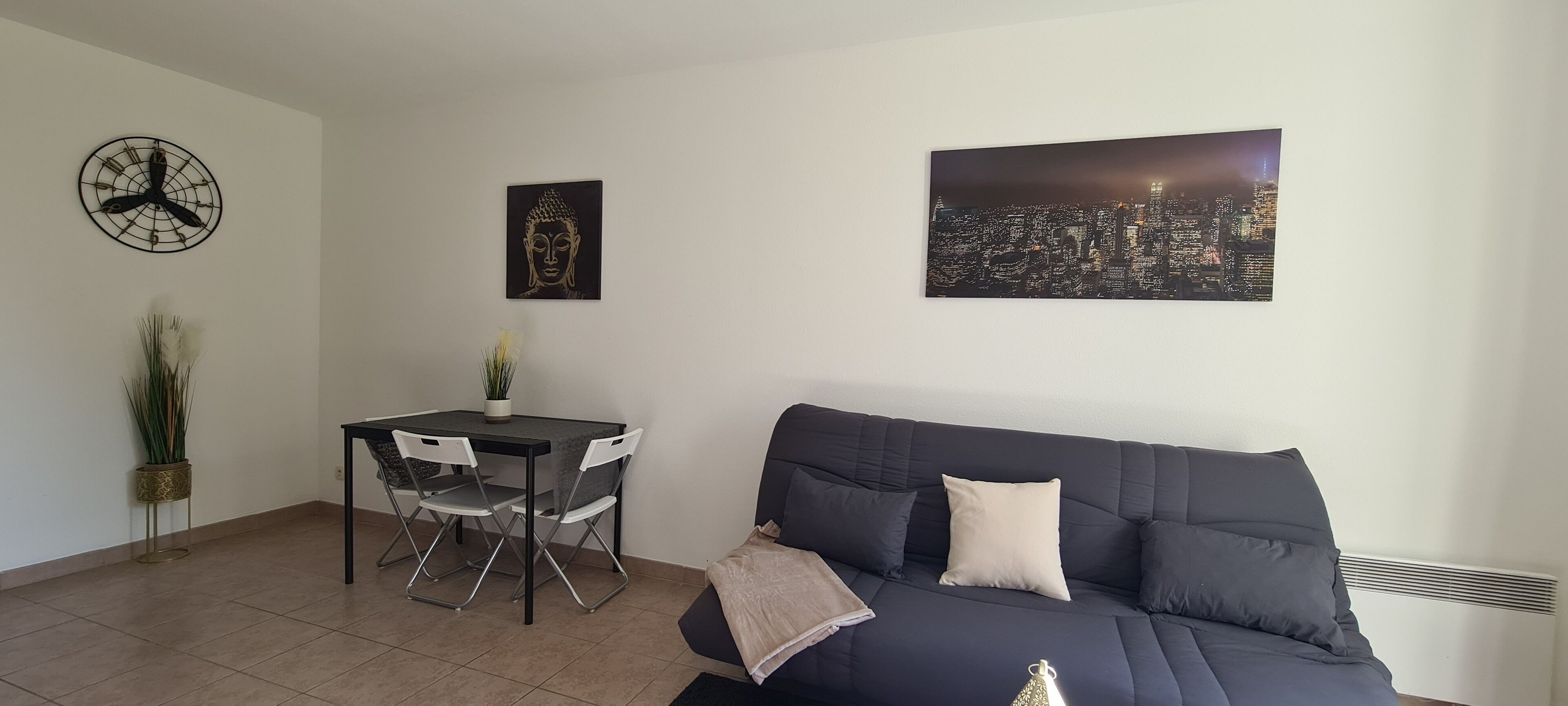 Ein Wohnzimmer mit einem dunkelblauen Sofa, einem Esstisch mit Stühlen, einer Wanduhr und zwei gerahmten Kunstwerken.