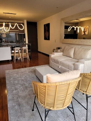 A modern living room with a sofa, two wicker chairs, a coffee table, and a kitchen area in the background.
