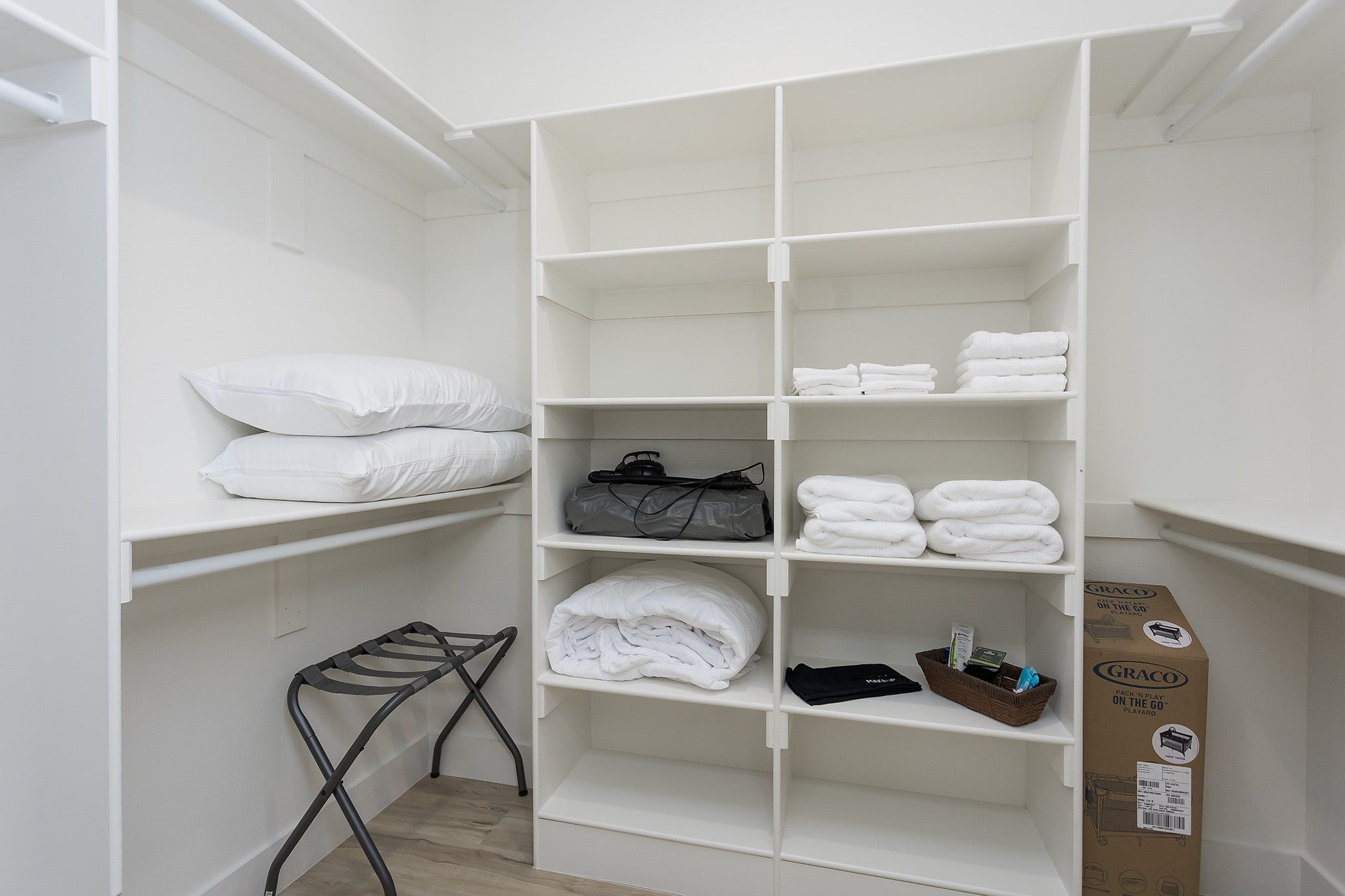 A white closet with shelves containing folded towels, a black bag, and a Graco pack n play playard.