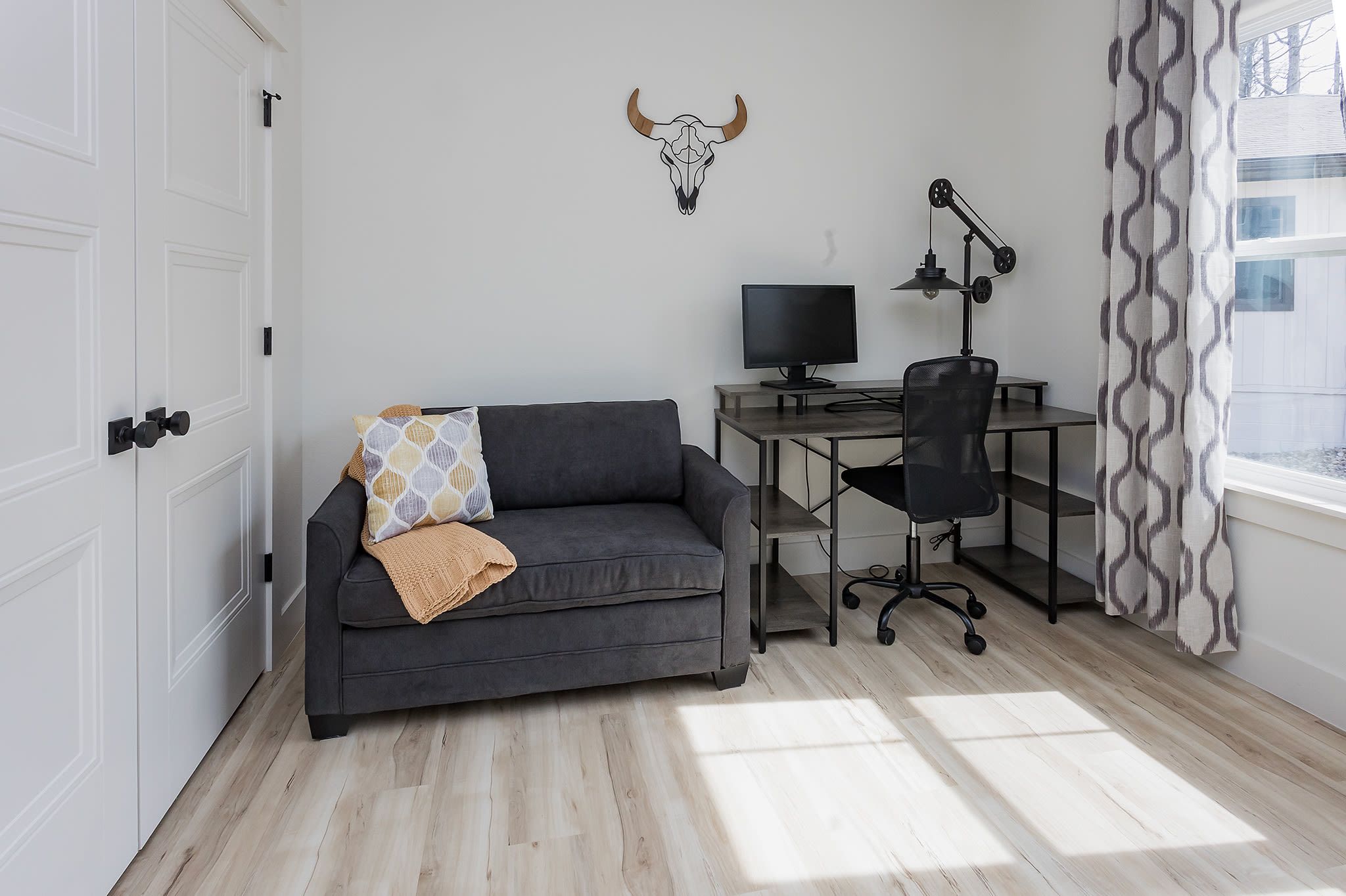 A modern living room with a grey sofa, a desk with a computer, and a decorative bull skull on the wall.