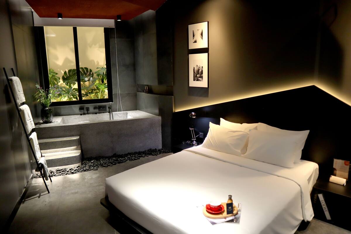 A modern hotel room with a large bed, a view of greenery through a window, a bathtub, and a small seating area with a towel and a plant.