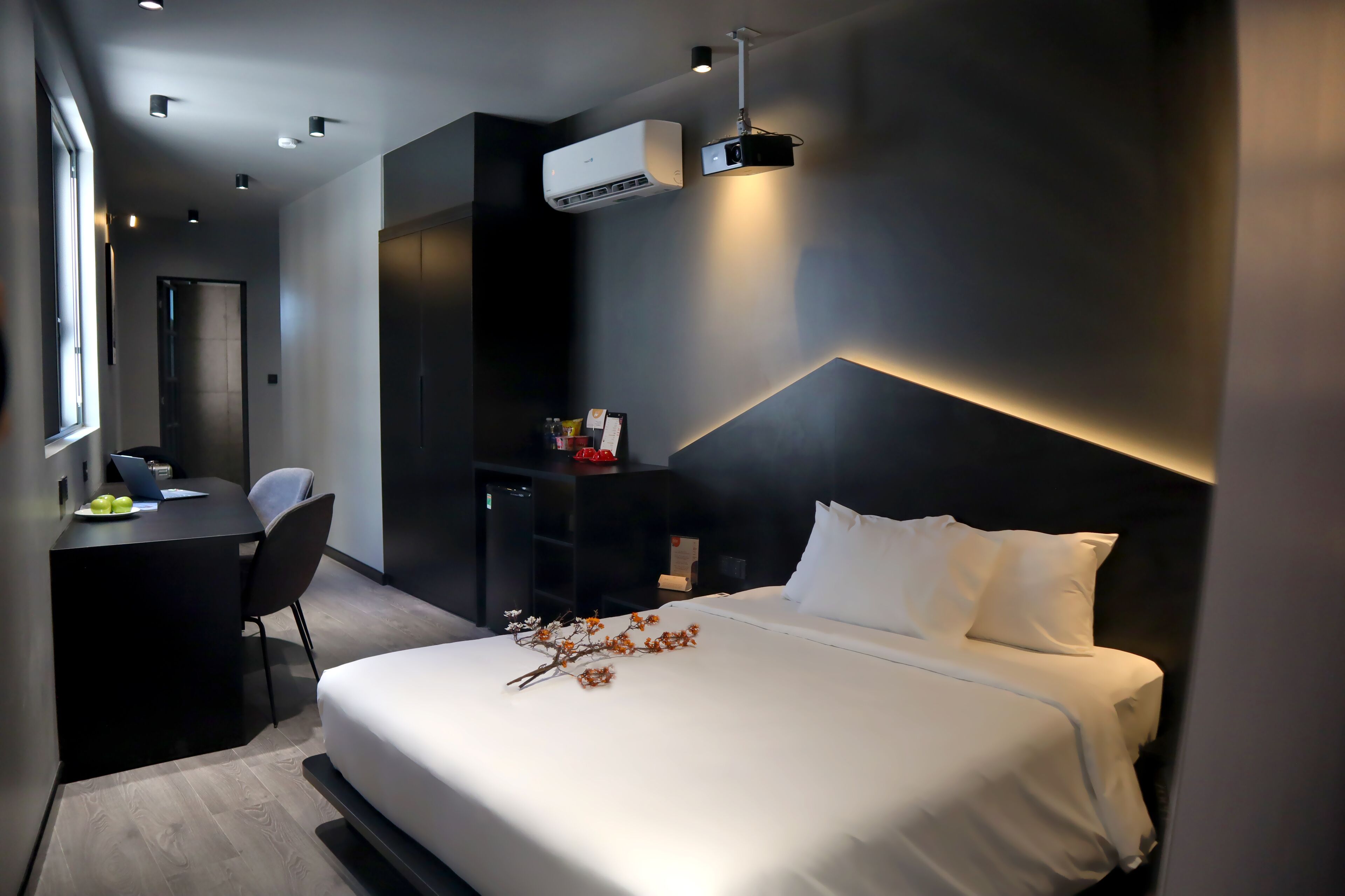 A modern hotel room with a large bed, a desk, and a chair. There is an air conditioning unit on the wall and a projector mounted on the ceiling.
