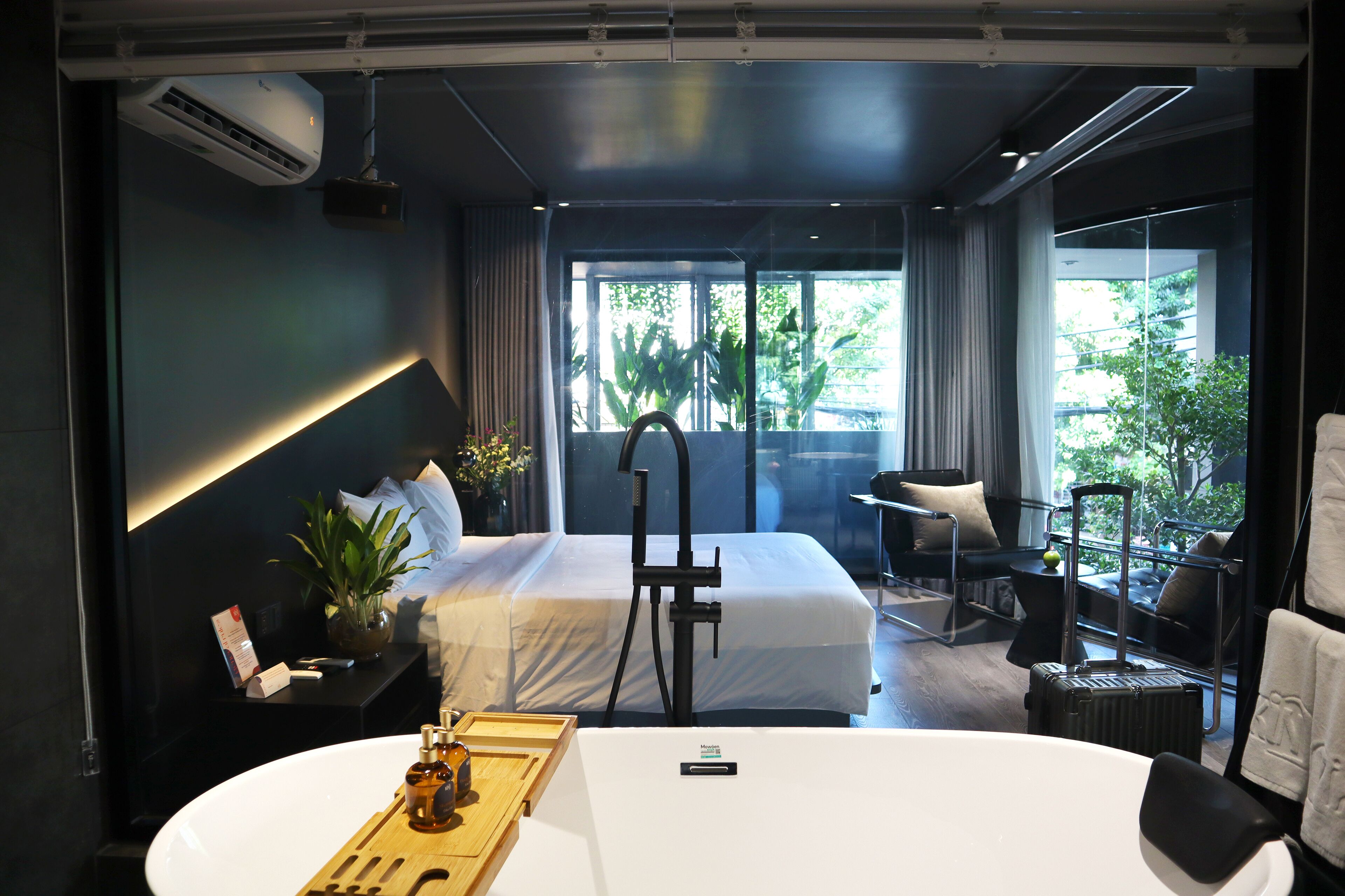 A modern hotel room with a large bed, a desk with a laptop, a suitcase, and a view of greenery through the window.