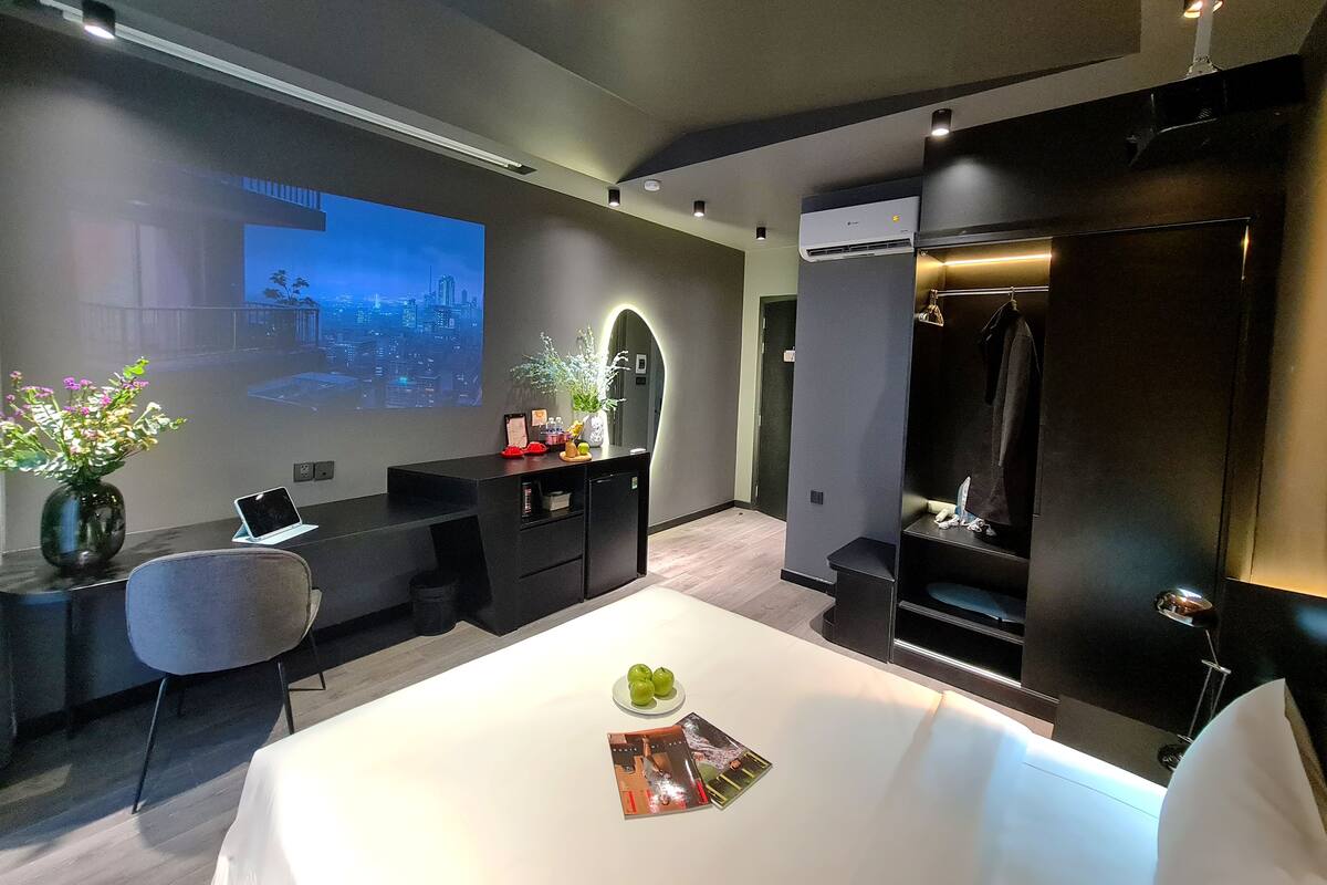 A modern hotel room with a large flat-screen TV displaying a cityscape, a desk with a laptop, a dining table with a magazine, and a black wardrobe.