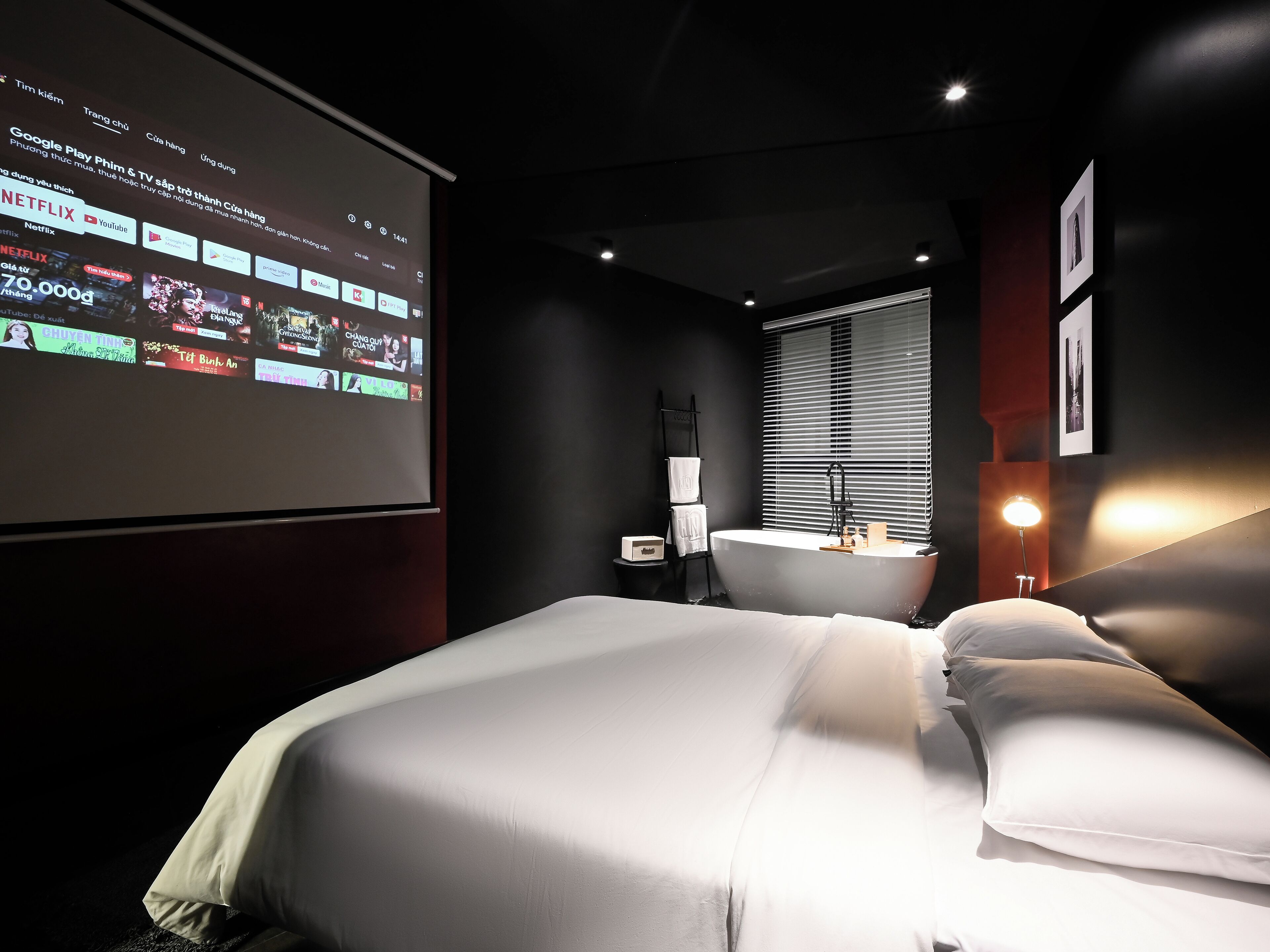 A hotel room with a bed, a desk, a bathroom, and a projector screen displaying a streaming service interface.