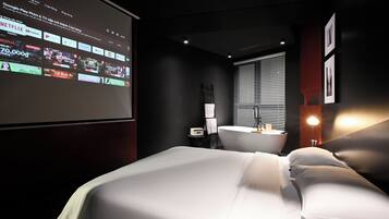 A hotel room with a bed, a desk, a bathroom, and a projector screen displaying a streaming service interface.