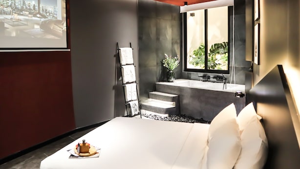 A modern hotel room with a large bed, a small bathroom, a window with a city view, and a projector screen displaying a cityscape. - Kin Wander Tan Binh, The Mountain (Ho Chi Minh City)