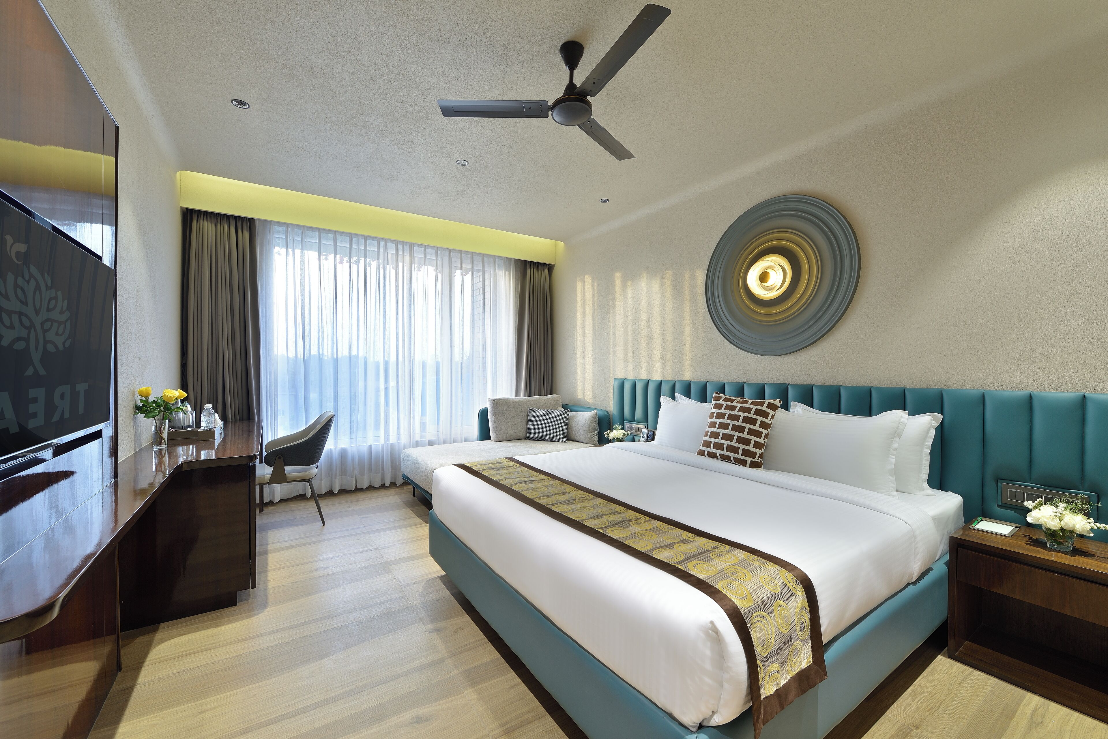 A hotel room with a large bed, a desk, a chair, a TV, and a ceiling fan.