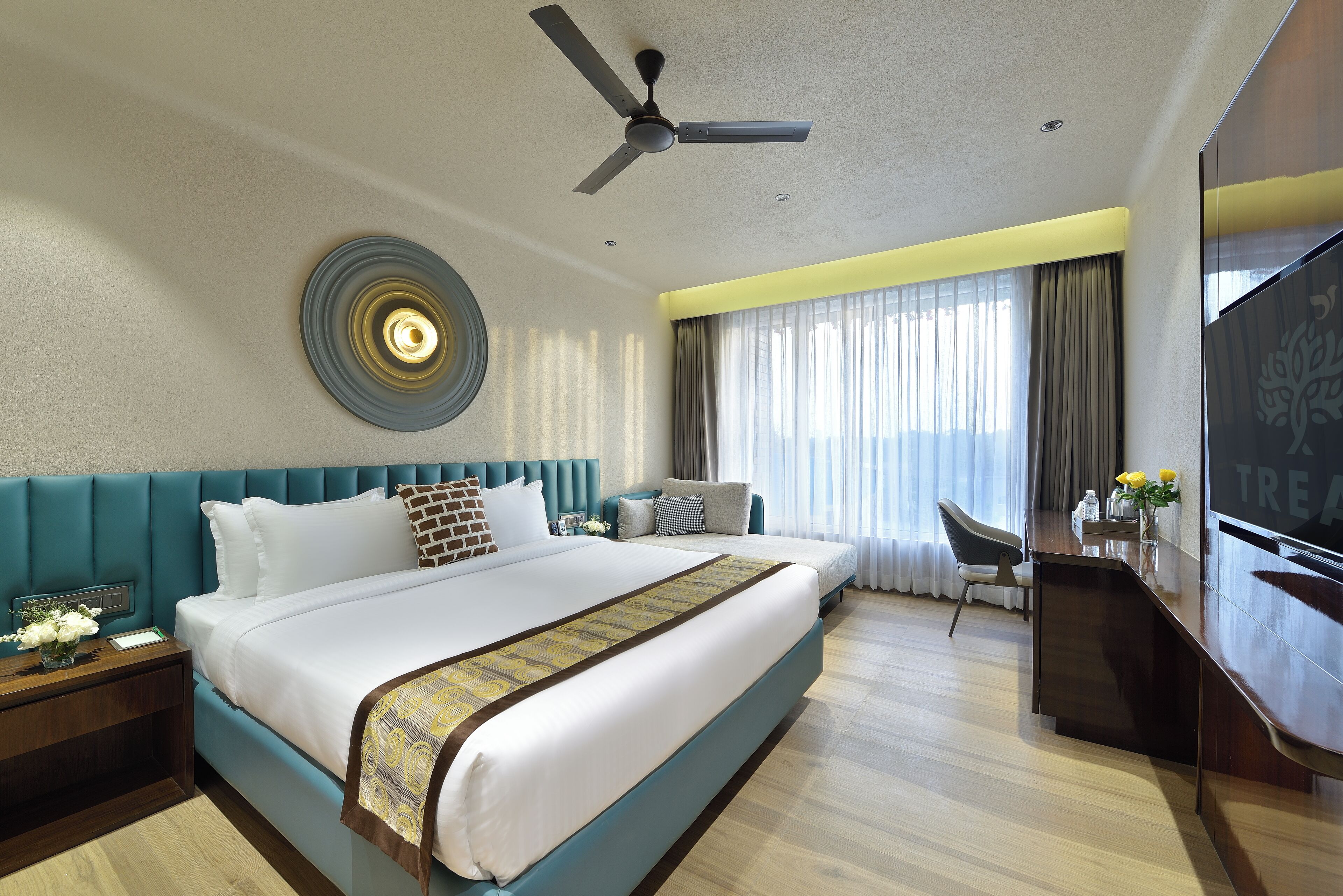 A hotel room with a large bed, a desk, a chair, a TV, and a ceiling fan.