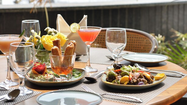 A table set with various dishes and drinks, a glass of water, and a glass of red drink, with a view of a water feature in the background. - URBANE BY TREAT  (Daman)