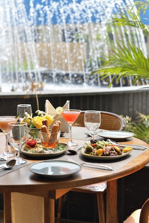 A table set with various dishes and drinks, a glass of water, and a glass of red drink, with a view of a water feature in the background.