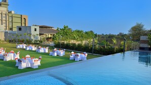 A poolside area with white tables and chairs, a clear blue pool, and a view of buildings and greenery. - URBANE BY TREAT  (Daman)