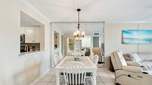 A modern kitchen with white cabinets, a dining area with a white table and chairs, and a living area with a beige sofa and a painting of a beach. - Oceanfront Resort 2BR Condo – Poolside, Scenic Pond Views, Sleeps 6 (St. Augustine)
