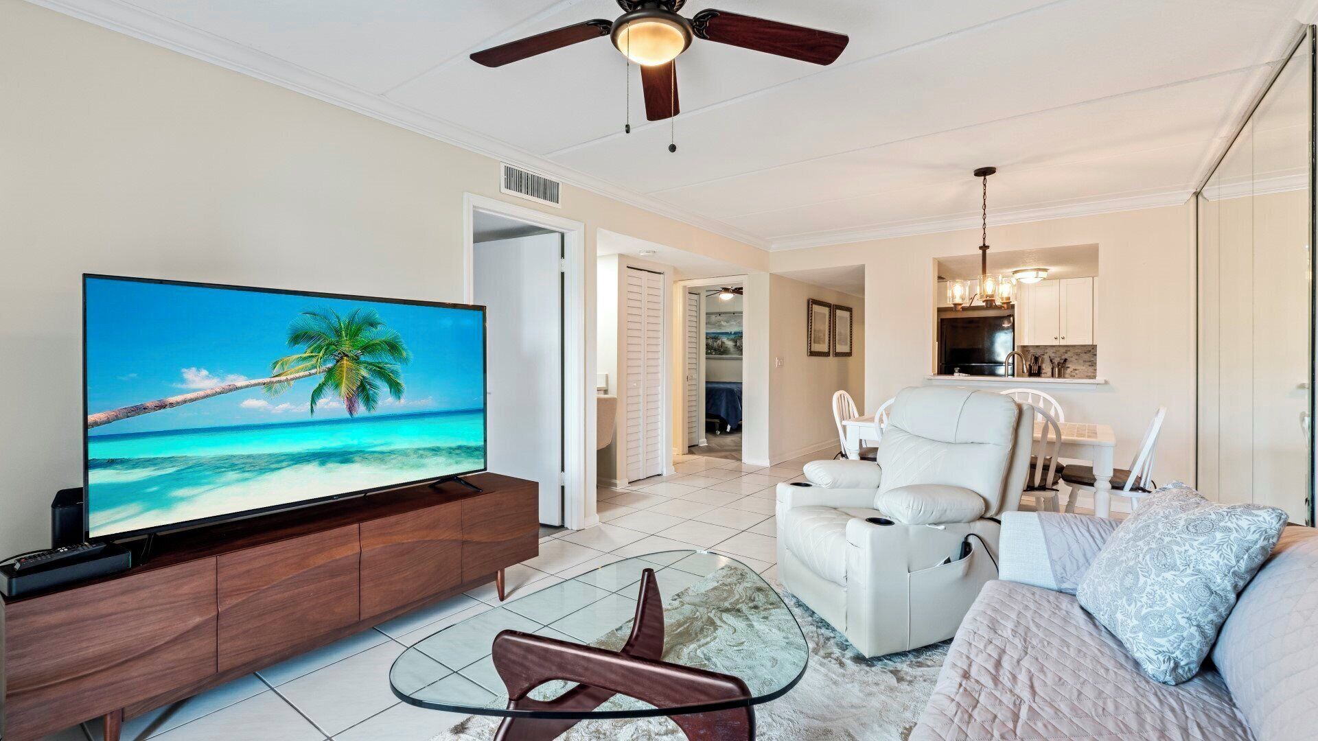 A living room with a flat-screen TV, a ceiling fan, a white sofa, and a glass coffee table.