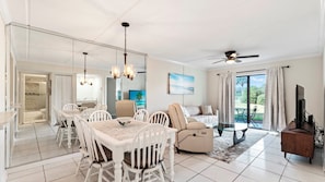 A spacious living area with a dining table, chairs, a sofa, a ceiling fan, and a large window with curtains.