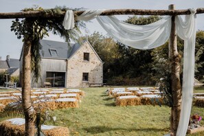 A rustic outdoor setting with a stone building, hay bales, and a wooden arbor with white fabric.