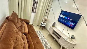 A living room with a brown sofa, a flat-screen TV mounted on the wall, a small white side table with a vase, and a window with curtains. - Apartamento a 4 min da Bas ílica  (Aparecida)