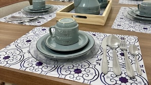 A dining table set with grey ceramic dishes and silverware, a wooden tray, and a potted plant in the background. - Apartamento a 4 min da Basílica  (Aparecida)
