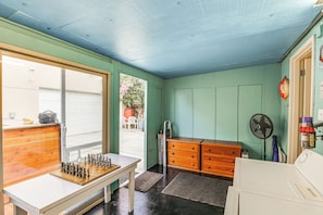 Quarto com um tabuleiro de xadrez sobre uma mesa, um ventilador e uma pequena cozinha com pia e armários.