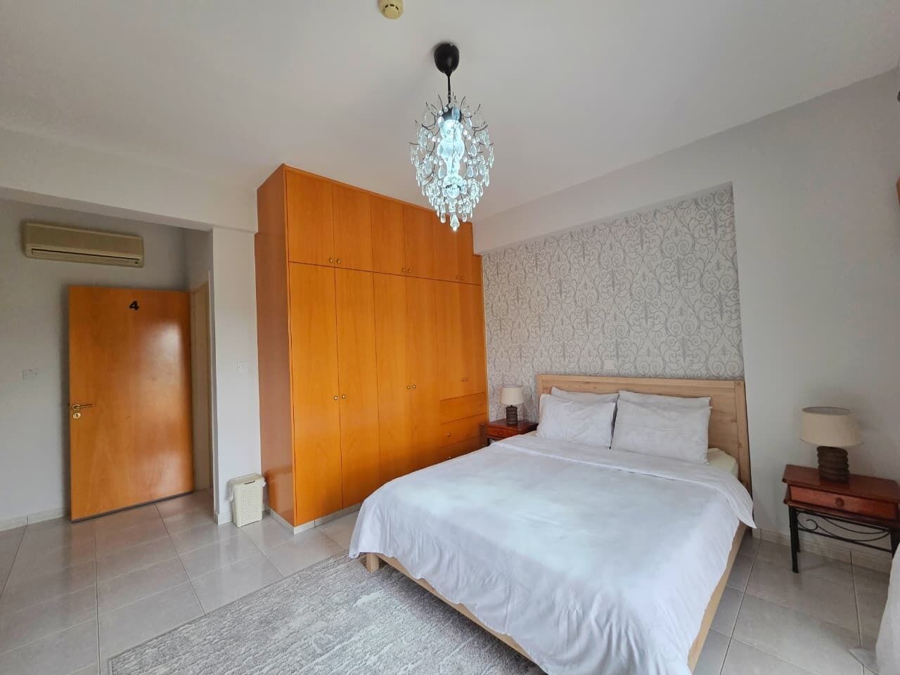 A hotel room with a large bed, wooden wardrobe, chandelier, and patterned wallpaper.