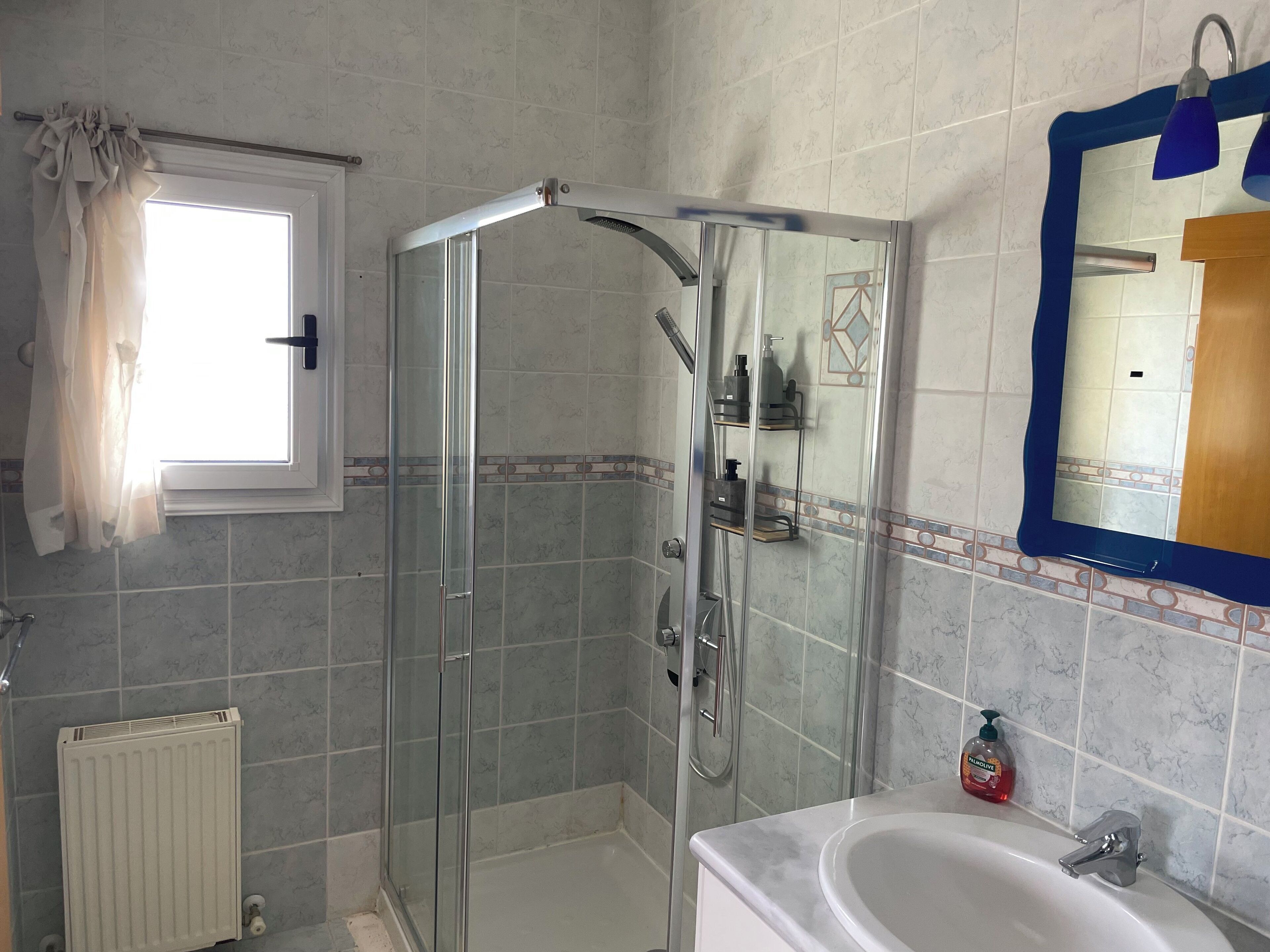 Un baño con ducha envuelta en vidrio, un lavabo blanco y un espejo con marco azul.
