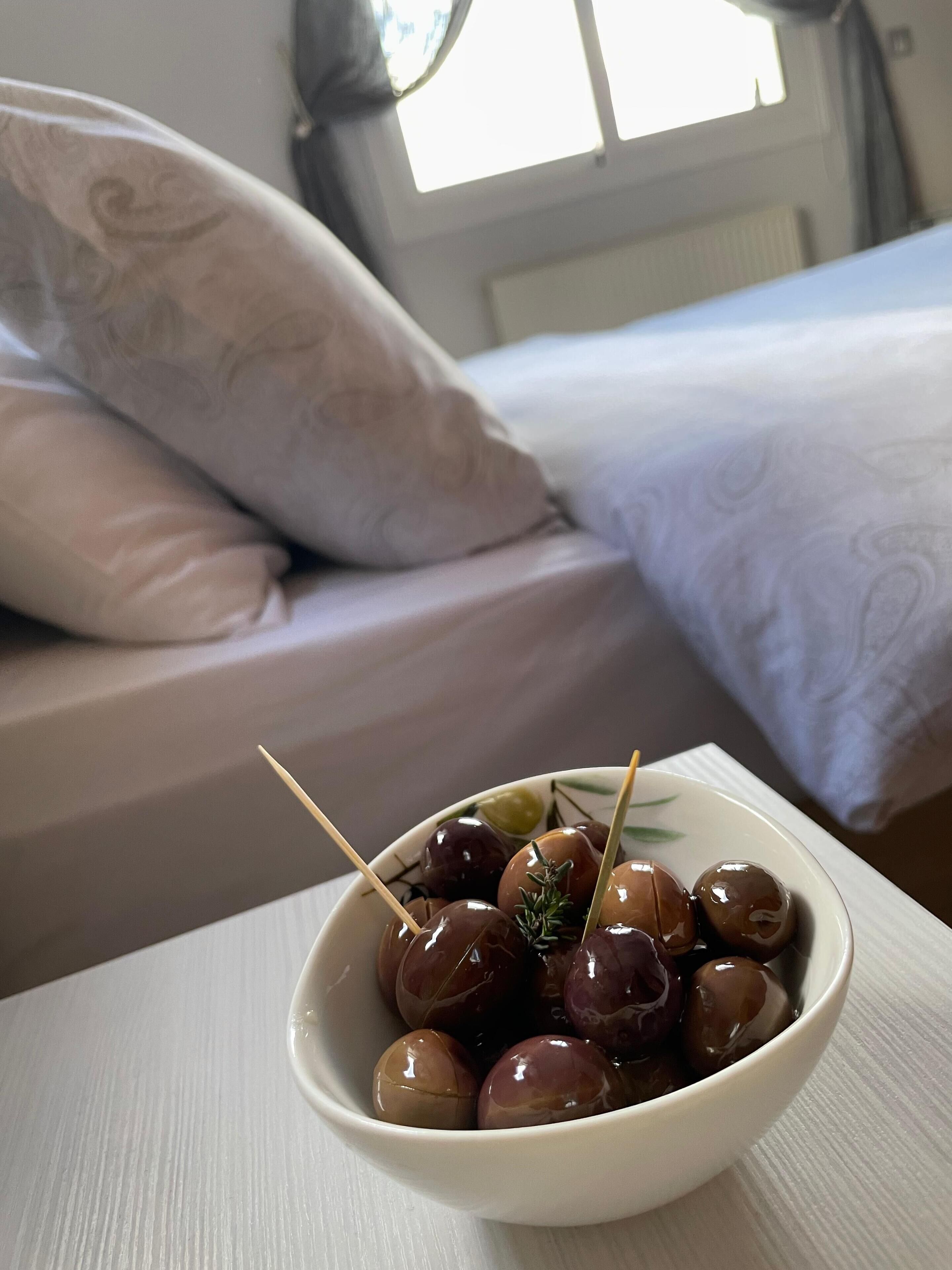 A bowl of cherries on a white table with a bed and pillows in the background.