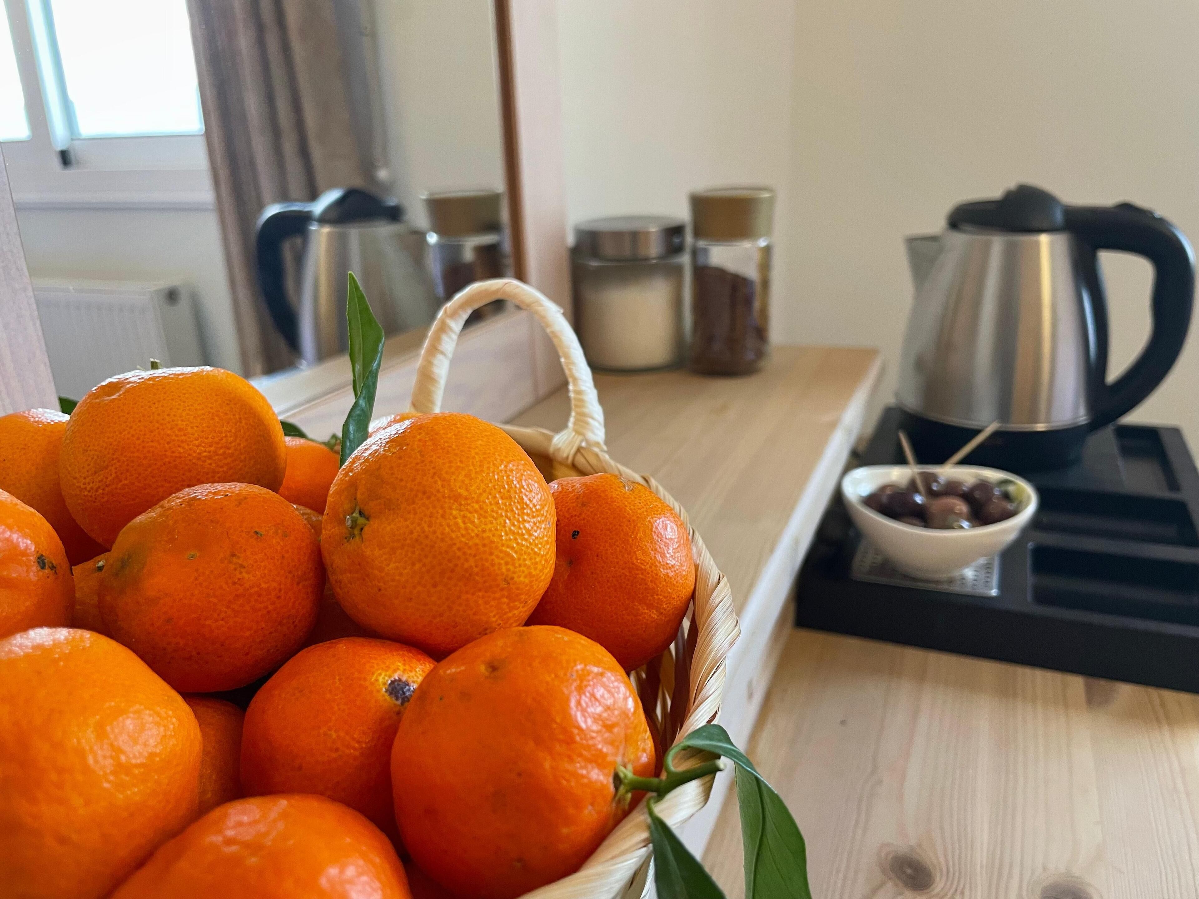 A basket of oranges on a wooden table with a kettle and a bowl of olives in the background.