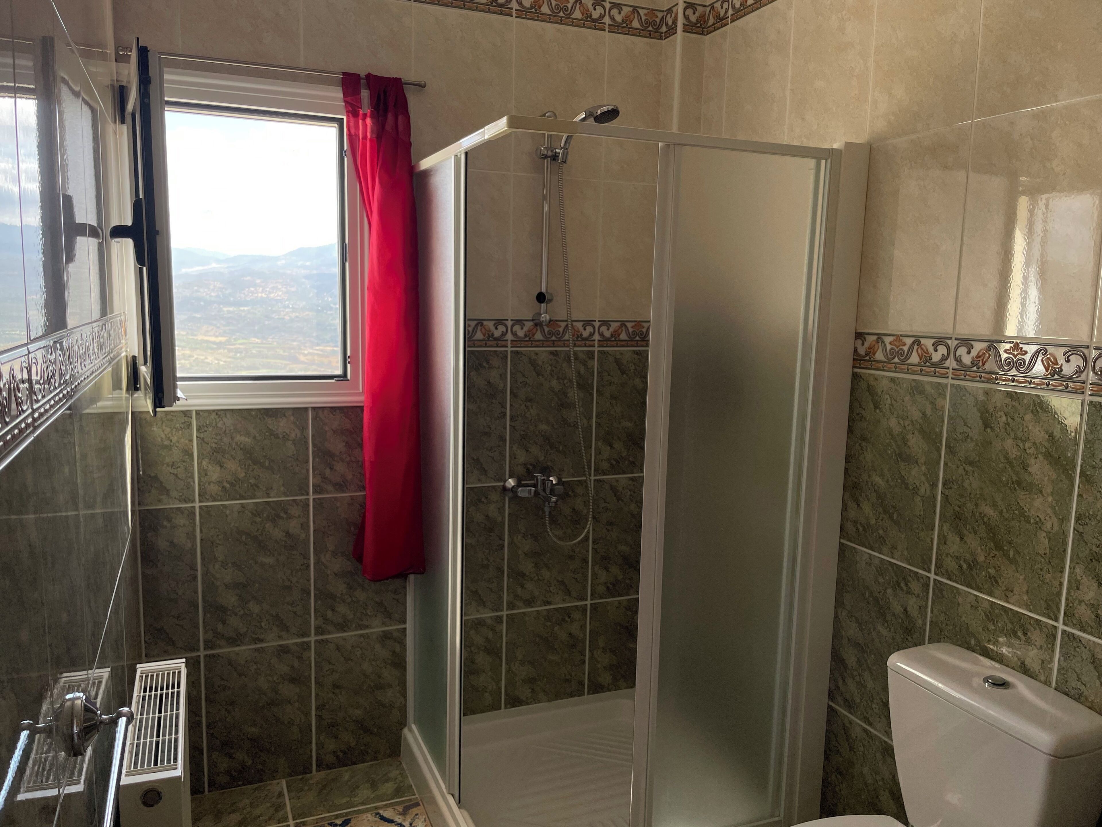 A bathroom with a glass-enclosed shower, a toilet, and a window with a red curtain.