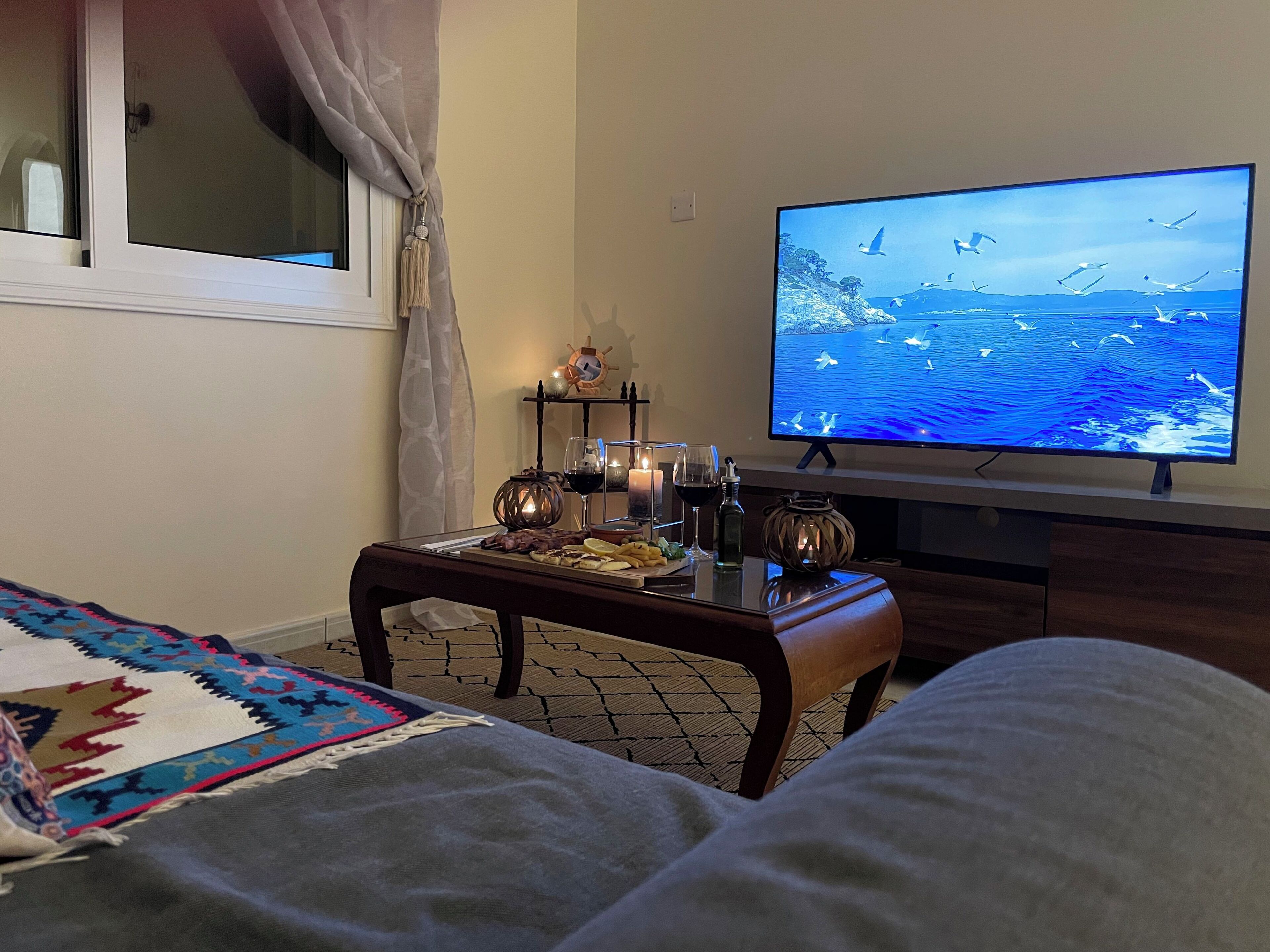 Una sala de estar acogedora con un televisor mostrando un paisaje marino, una mesita con aperitivos y bebidas, una alfombra estampada y una ventana con cortinas.