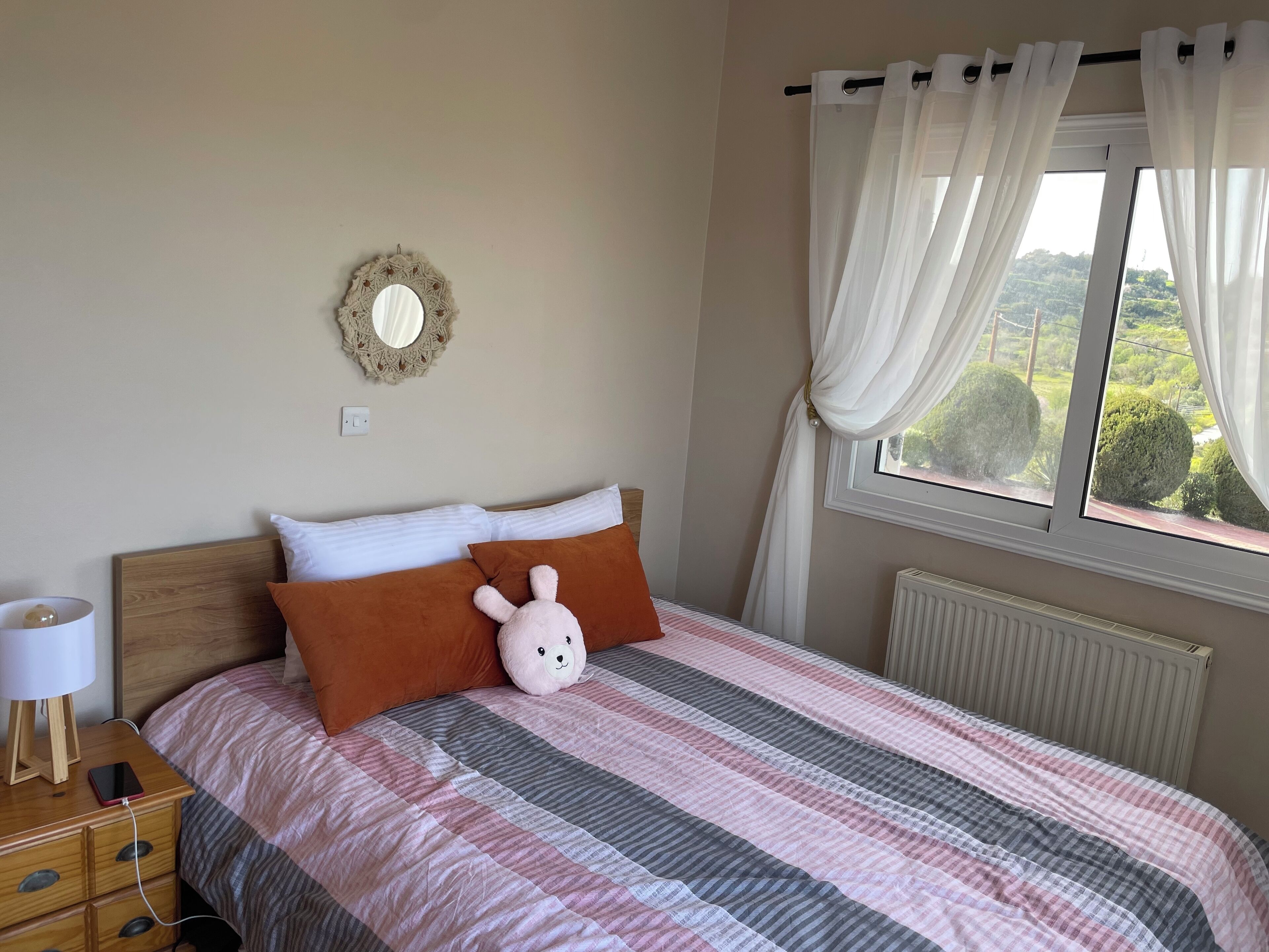 Quarto com cama, mesa de cabeceira com abajur, espelho e janela com cortinas.