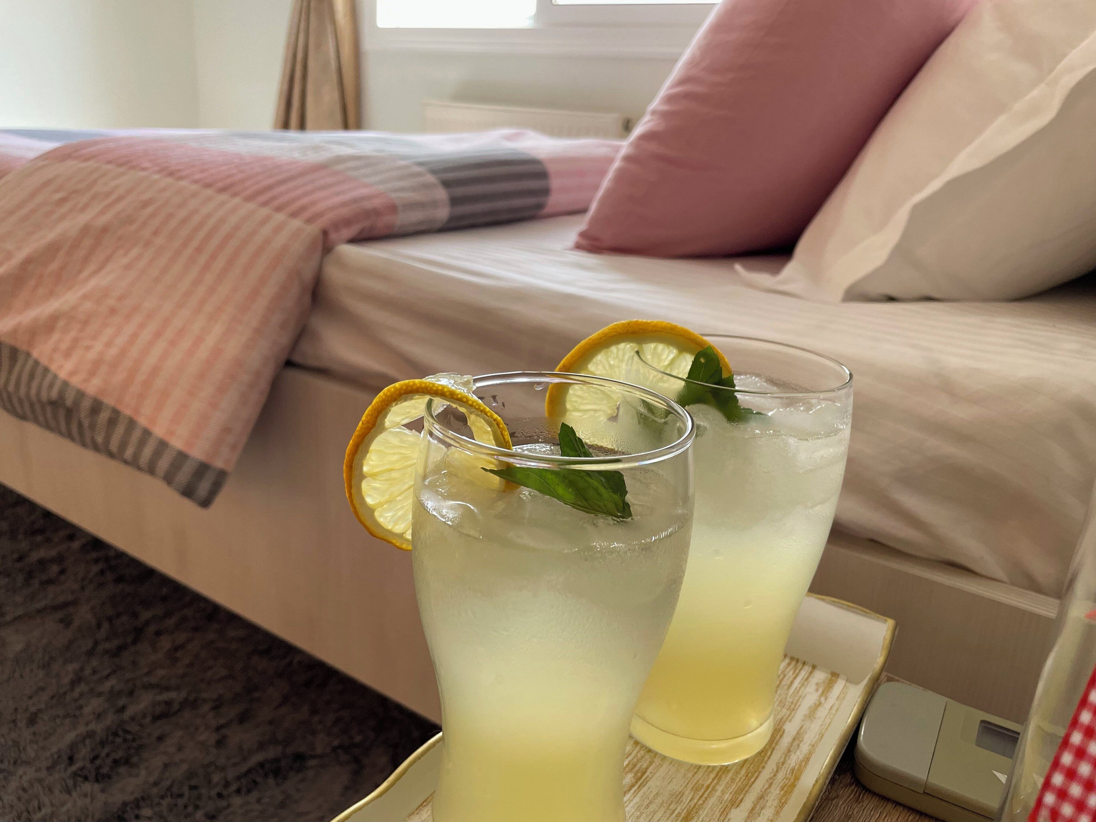 Dos vasos de bebida helada con rodajas de limón y hojas de menta sobre una bandeja, colocados sobre una cama con una manta rosa y almohadas.
