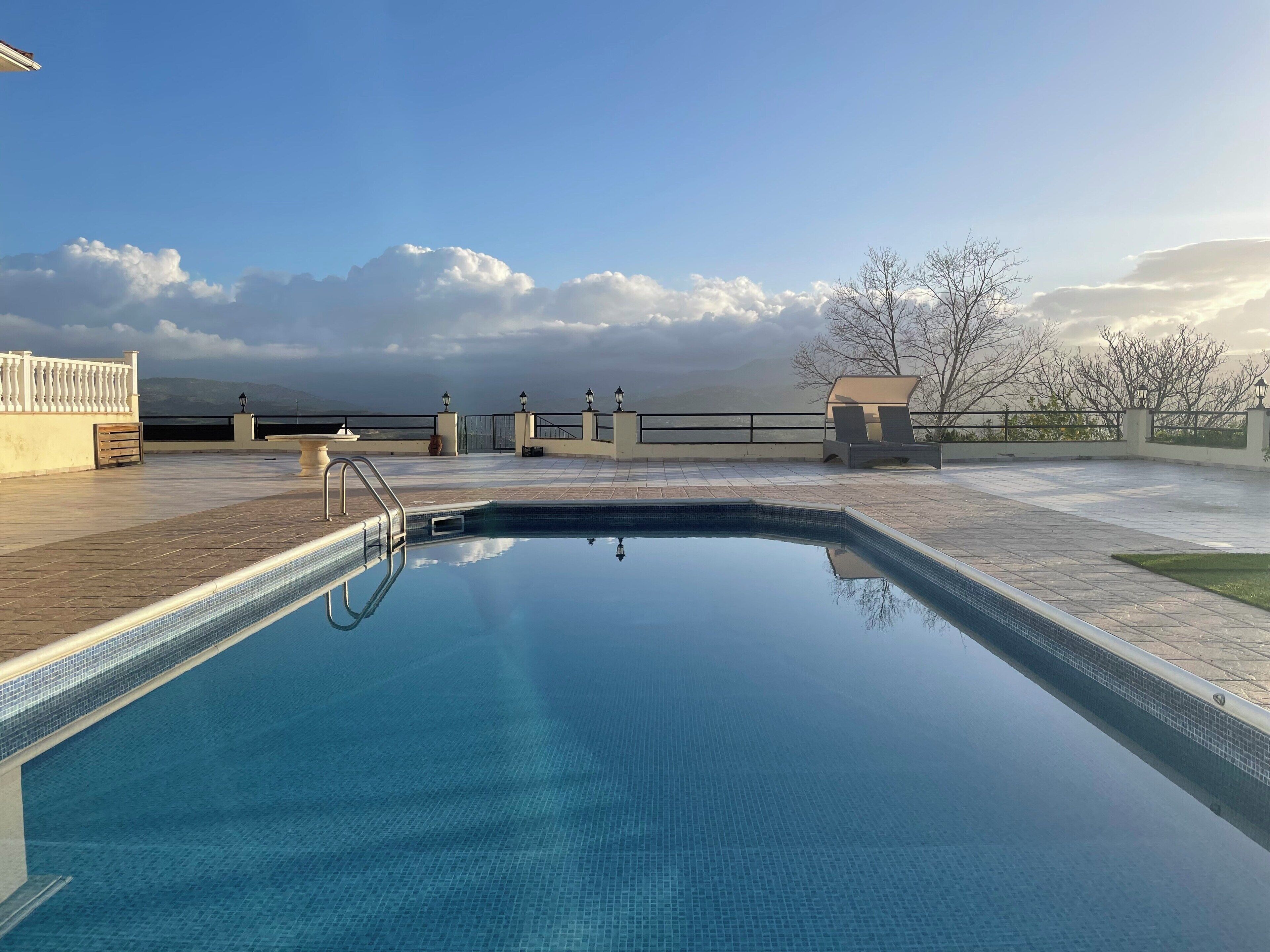 Piscina azul cristalina con borde de azulejos, rodeada por una zona de patio con barandilla y vistas al cielo y montañas.