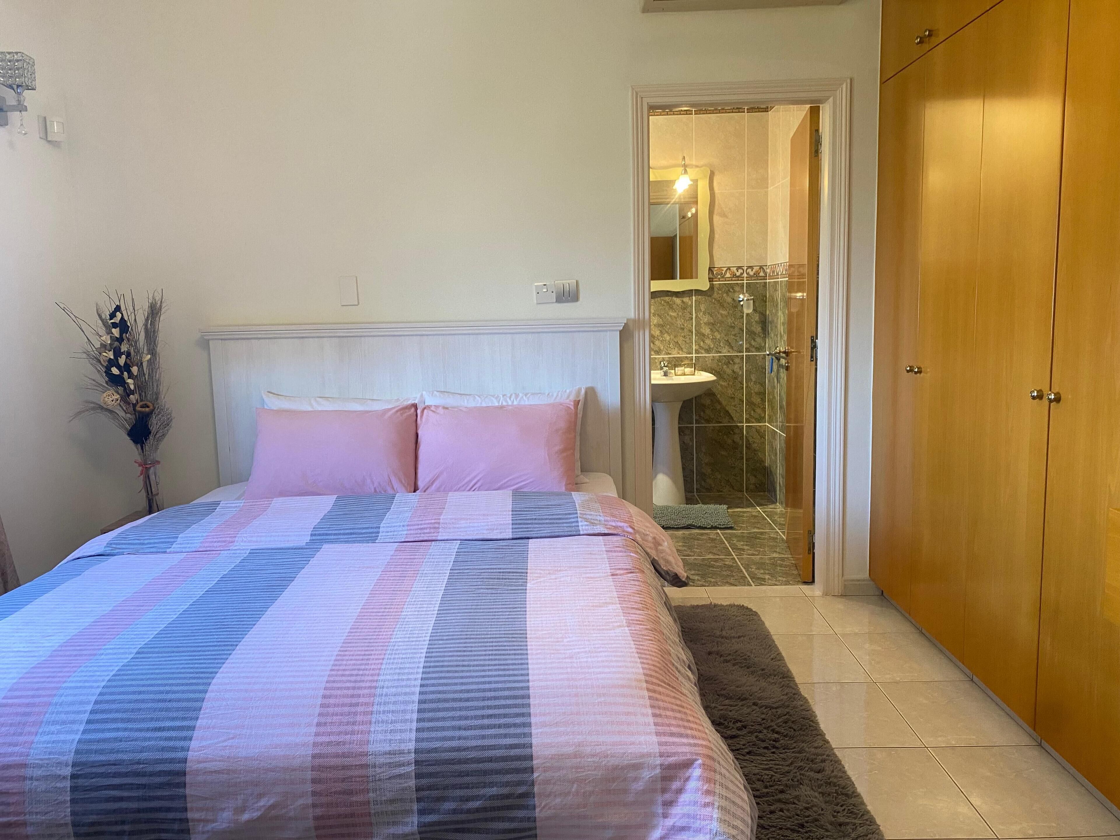 Un dormitorio con cama, mesita de noche, espejo, armario y baño visible a través de una puerta abierta.