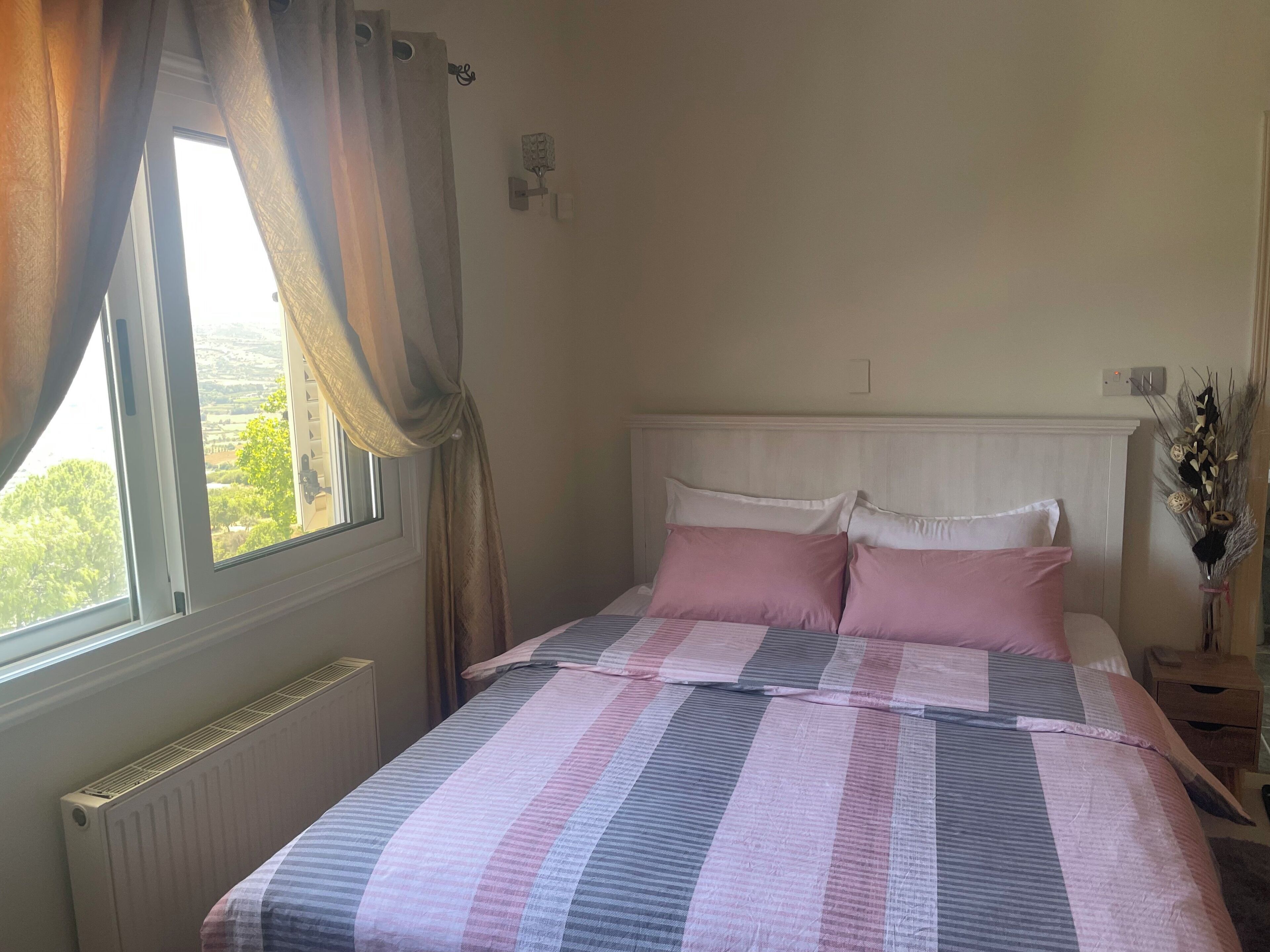 A bedroom with a bed, curtains, a window, a radiator, and a nightstand with a vase.