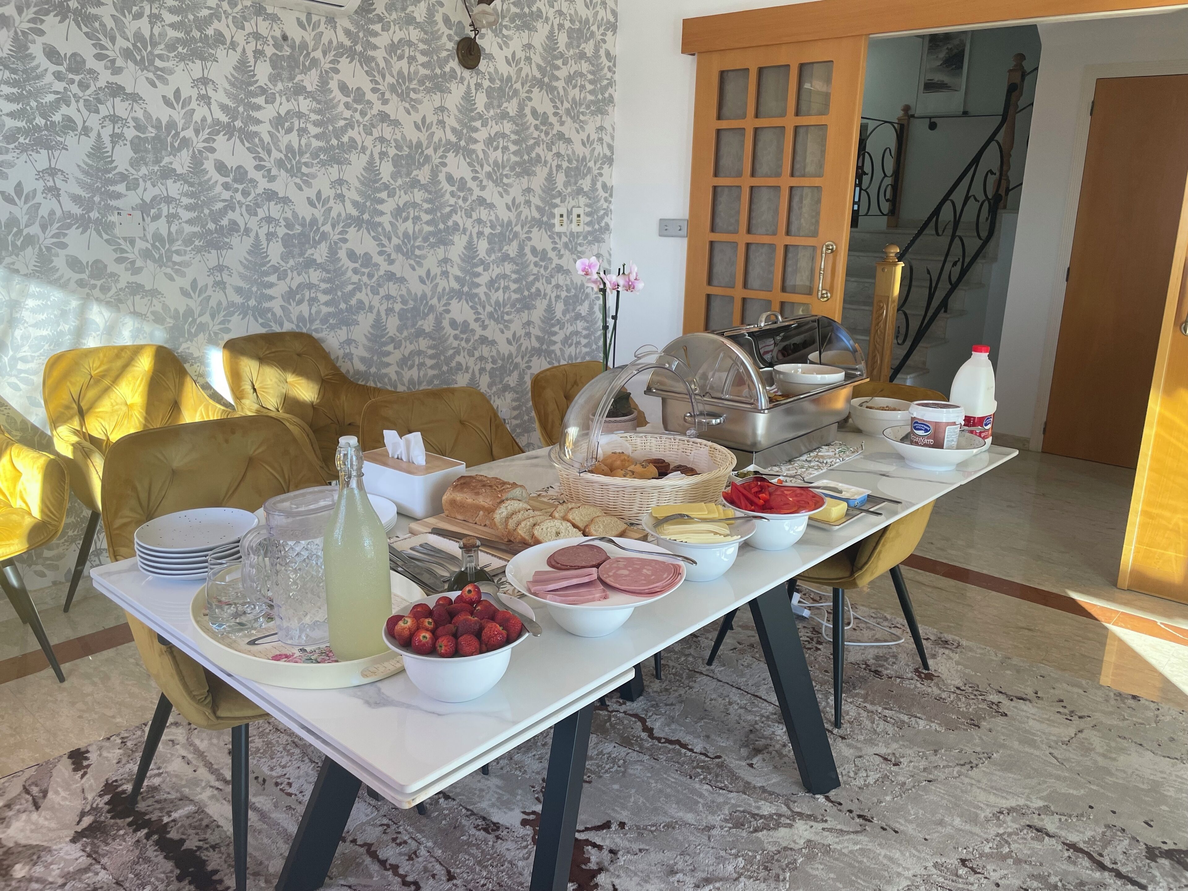 Un tavolo imbandito con diversi cibi, un buffet e sedie gialle in una stanza con carta da parati decorata e una scala sullo sfondo.
