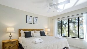 Ein Schlafzimmer mit einem großen Bett, zwei Nachttischlampen, einem Deckenventilator und einem Fenster mit Blick auf Bäume.