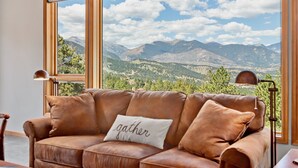 A brown leather sofa with a cushion that says "gather" in front of a large window with a mountain view. - Aspen Spirit Vacation Home At Windcliff 3 Bedroom Home by RedAwning (Estes Park)