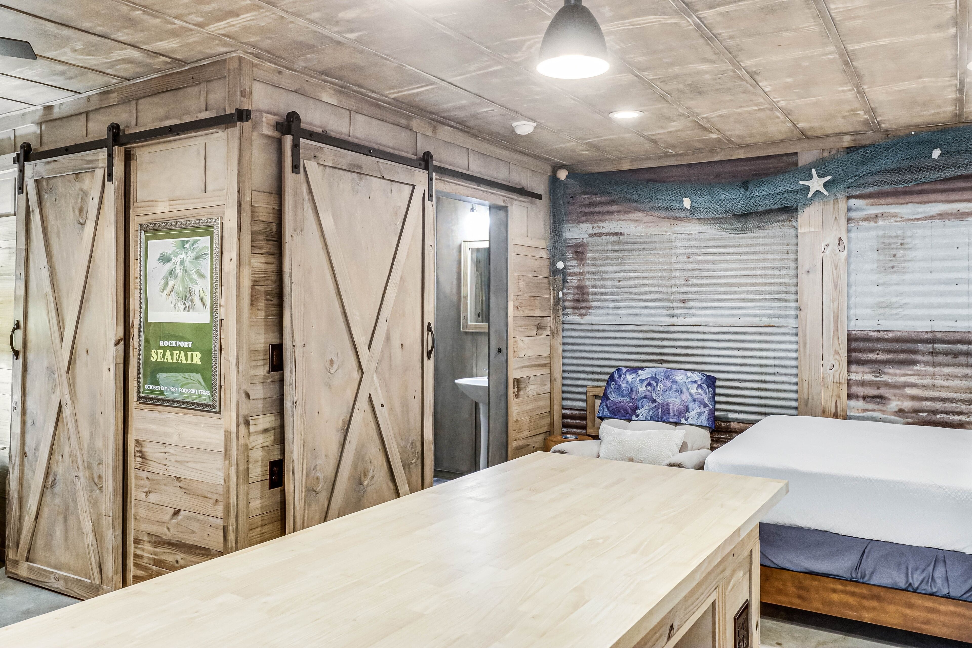 Una habitación con una mesa de madera, una cama, un baño con un lavabo y una puerta con un cartel que anuncia una feria marítima.